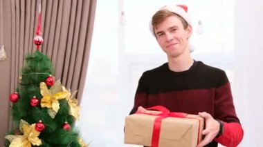 Noel Baba şapkalı genç beyaz adam Noel ağacının yanında duran kırmızı kurdeleli hediyeyi veriyor..