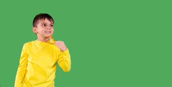 Foto de um menino de idade escolar sorridente com capuz amarelo olhando para o lado e segurando o brinquedo de dinossauro em seu ombro contra o fundo verde, espaço vazio. — Fotografia de Stock