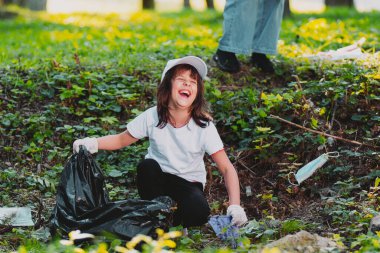Elinde çöp torbası olan ve plastik çöp toplayan, beyaz eldivenlerle yerde oturan gülen genç bir kızın yakın plan fotoğrafını çek..