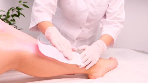 Cosmetólogo en guantes médicos en un salón de belleza hace procedimiento de depilación de las piernas de la mujer usando cera y tiras. — Vídeos de Stock