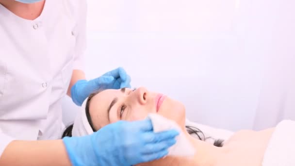 Cosmetóloga profesional en máscara médica y guantes limpia su cara de mujer paciente con una servilleta después de un procedimiento cosmético para rejuvenecer la piel de la cara. — Vídeos de Stock