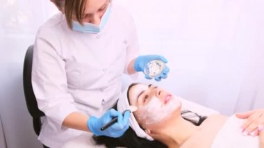 Tıbbi maskeli ve silikon fırçalı eldivenli bir güzellik uzmanı spa güzellik salonundaki genç bir bayan müşterinin yüzüne beyaz nemlendirici bir yüz maskesi takıyor..