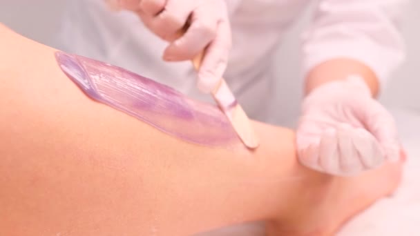 Esteticista profesional con guantes blancos y uniforme aplica cera usando palo de madera para depilar las piernas de las mujeres jóvenes en el salón de cosmetología. — Vídeos de Stock