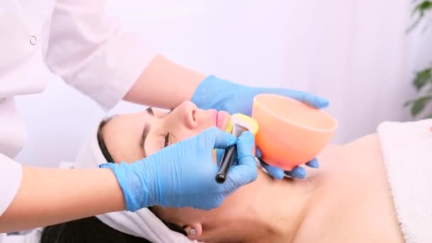 La esteticista profesional que usa guantes azules médicos con un cepillo aplica una máscara hidratante amarilla a la cara de una mujer joven en un salón de belleza de spa. — Vídeo de stock