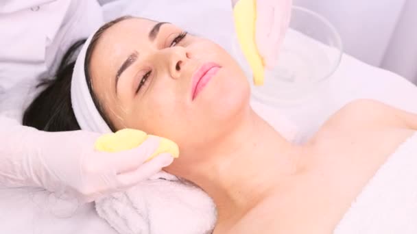 Молодая красивая женщина расслабляется, в то время как профессиональный косметолог очищает лицо двумя губками, готовясь к процедурам омоложения лица. — стоковое видео