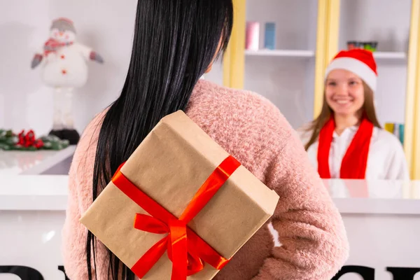 Arkadaki fotoğrafta, Noel Baba şapkalı ve kırmızı eşarplı meslektaşına vermek için arkasında kırmızı kurdele olan hediye kutusunu tutan esmer bir kadın görülüyor.. — Stok fotoğraf