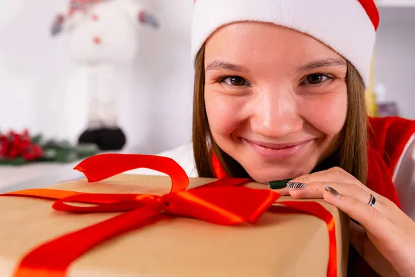 Снимок красивой девушки в шляпе Санта-Клауса трогает подарочную коробку подбородком и рукой с цветным маникюром. — стоковое фото
