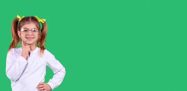 Jeune fille intelligente portant l'uniforme de l'école et des lunettes debout sur un fond vert avec expression faciale heureuse et tenant son index sur sa joue, espace de copie. — Photo