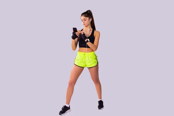 A pose comprimento de uma menina fitness bonita magro mantendo sua uma perna para a frente usando roupas esportivas usando telefone inteligente, relógio digital e fones de ouvido. — Fotografia de Stock