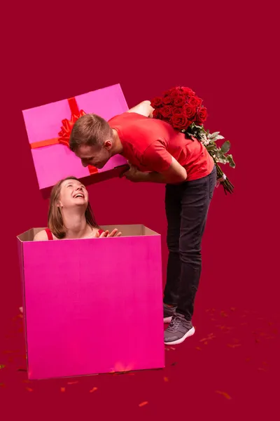 Elinde güzel bir buket kırmızı gül tutan bir erkek arkadaşın resmi büyük bir hediye kutusunu açıyor ve içinde kız arkadaşını görüyor.. — Stok fotoğraf