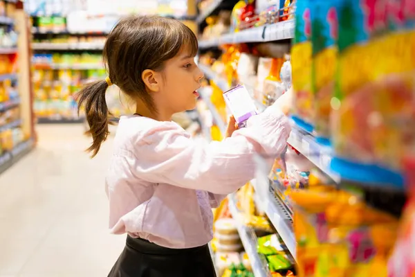 Σχολείο κορίτσι στο σούπερ μάρκετ ψάχνει για το προϊόν που της αρέσει στα ράφια και προσπαθεί να επιλέξει από τη μεγάλη ποικιλία των πολύχρωμων προϊόντων. — Φωτογραφία Αρχείου