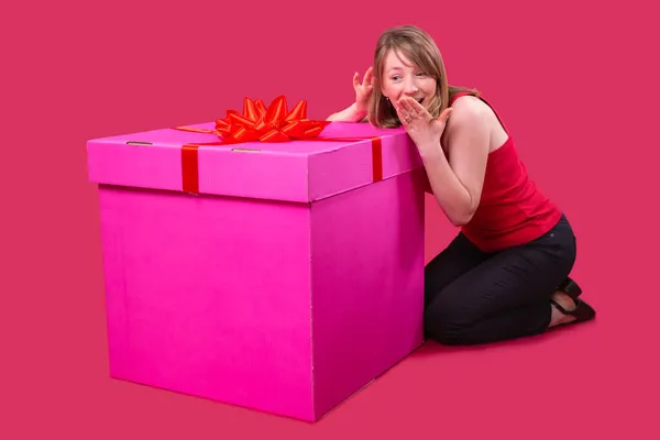 Kırmızı tişörtlü ve siyah kotlu genç bir bayanın fotoğrafını çek. Pembe renkli büyük hediye kutusunun yanında oturuyor ve ağzını eliyle kapatıyor. Şaşırmış yüz ifadesi var.. — Stok fotoğraf