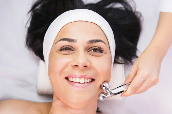Foto ravvicinata di una donna cliente felice in un salone di bellezza, cosmetologo professionista che fa il suo massaggio rilassante del viso con rullo cosmetico. — Foto Stock
