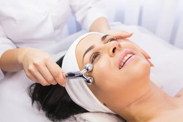 Jovem relaxada sofre o procedimento de massagem facial de rejuvenescimento em um salão de beleza por um cosmetologista usando rolo facial. — Fotografia de Stock