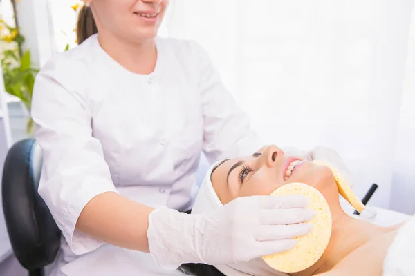 Cosmetologo professionista che effettua la procedura di pulizia per idratare il viso con due spugne a una donna in un salone di bellezza. — Foto Stock