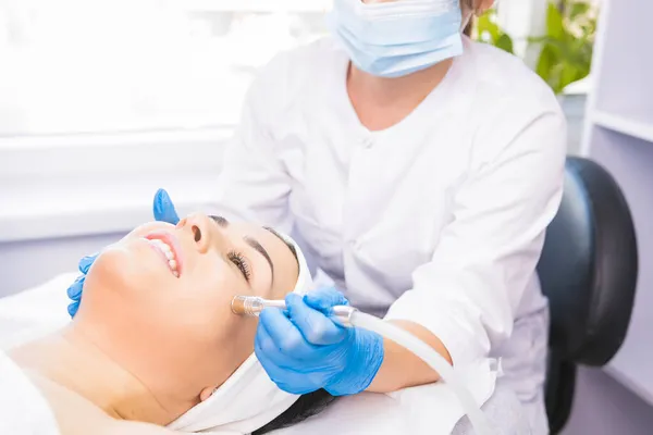 Kosmetikerin in einer medizinischen Schutzmaske und Handschuhen, die ihrer Klientin in einem Schönheitssalon die Mikrodermabrasion des Gesichts ermöglicht. — Stockfoto