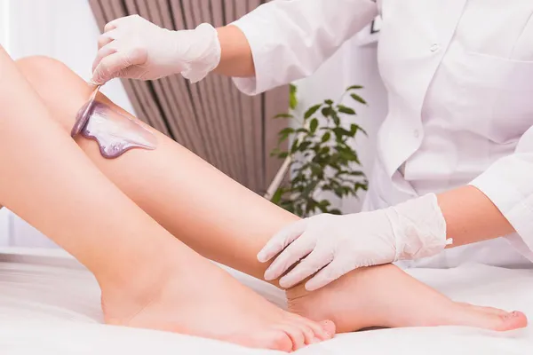 A beauty master in protective gloves applies violet depilatory pearl wax to a young womans leg for hair removal, beauty concept. — Stock Photo, Image