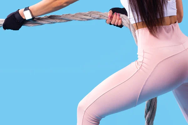 Η φωτογραφία πίσω όψη των γοφών ενός νεαρού λεπτού κοριτσιού με μακριά μαλλιά σε ροζ αθλητικά κολάν και d λευκό καπάκι κάνει ασκήσεις χρησιμοποιώντας το σχοινί fitness, τραβάει το σχοινί προς το μέρος του εαυτού της. — Φωτογραφία Αρχείου
