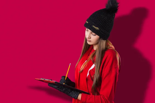 Foto vista lateral de una chica bonita vestida con ropa roja, sombrero negro y guantes sosteniendo el documento en sus manos y escribiendo con un lápiz. —  Fotos de Stock