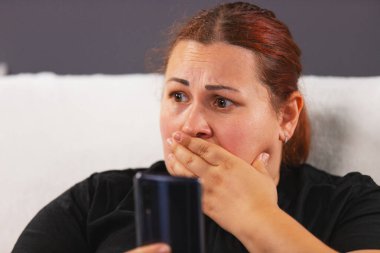 Siyah tişörtlü, endişeli bir kadının elinde akıllı bir telefonla beyaz koltukta oturduğu ve TV 'de kötü haberleri izlerken eliyle ağzını kapattığı bir fotoğraf..