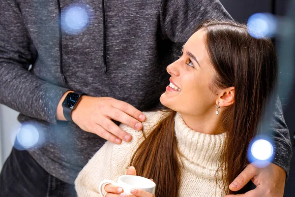 Szczęśliwa uśmiechnięta młoda dama w białym ciepłym swetrze trzymająca filiżankę herbaty w dłoniach patrząc na swojego chłopaka przytulającego jej ramiona. — Zdjęcie stockowe