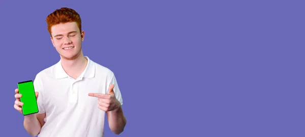 Young red hair man teenager in white t-shirt holding the smartphone in one hand and with the other pointing at the phone with green screen standing on violet background, cope space. — Stock Photo, Image
