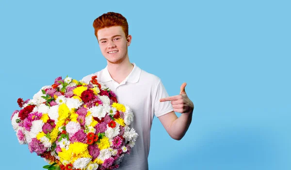 幸せな若いです男を保持している巨大な美しい花束の花とそれを指しています彼の指上に青の背景. — ストック写真