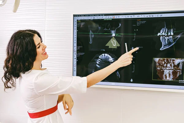 Beyaz üniformalı ve kırmızı kemerli güzel dişçi dişçi dişçi muayenehanesindeki ekranda dişlerinin fotoğrafını gösteriyor..