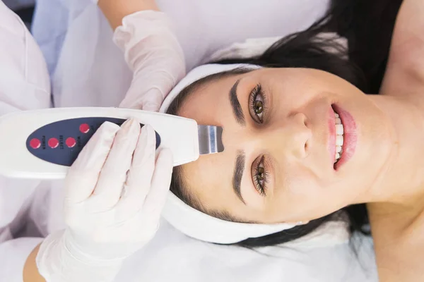Photo from above of a beautiful happy woman smiling passing the rejuvenation face procedure in beauty clinic and cosmetologist using Ultrasonic skin equipment. — Stockfoto