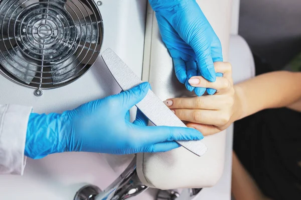 Manicurist filing young woman nails in salon wearing blue protective gloves make nails ready to polish. — Stockfoto