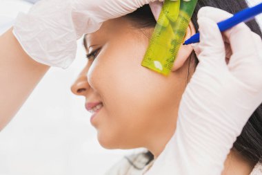 Kozmetik uzmanı elinde cetvel ve kalemle sabırlı bir kadının kulağının küpe deliğini ölçer..