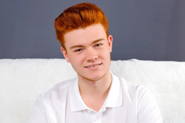Close up photo of a young red hair man in white t-shirt sitting on a sofa over violet background posing for a camera. — Stockfoto