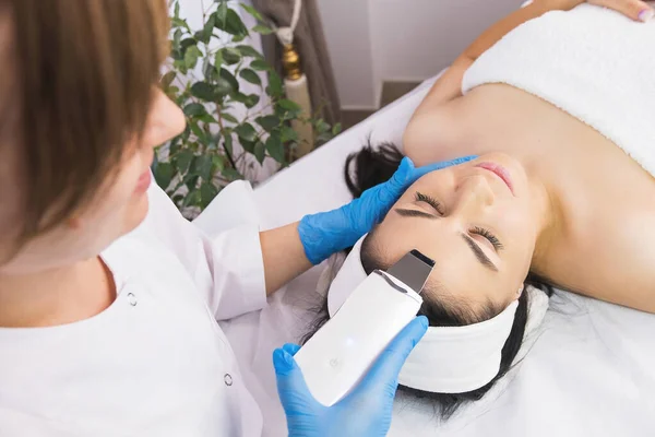 Specialist cosmetologist making facial procedure treatment to a patient woman using ultrasonic skin equipment. — Stock Fotó
