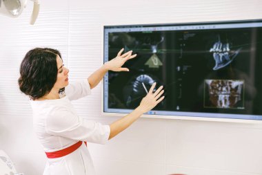 Serious dentist woman in white uniform and red belt showing attentively at the monitor with teeth snapshot.