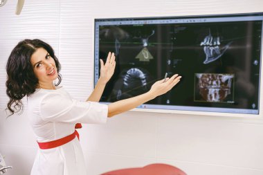 Stomatologist brunette woman with smile in white uniform holding her hands widely and showing at the monitor with teeth snapshot.