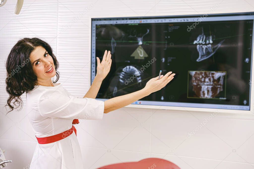 Stomatologist brunette woman with smile in white uniform holding her hands widely and showing at the monitor with teeth snapshot.