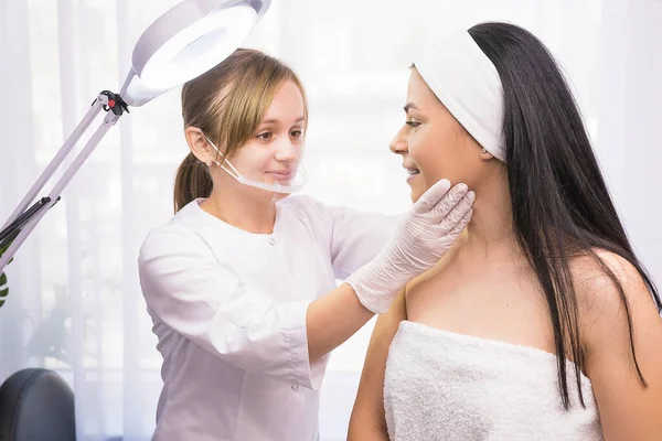 Kosmetologe in Uniform, medizinischer Kunststoff-Gesichtsmaske und Handschuhen, der eine Patientin in einem Klinik-Wellness-Salon untersucht. — Stockfoto