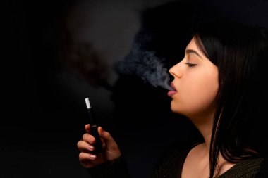 Genç bir kadın tüttürüyor ve koyu bir arka planda sigara dışarı salıyor..