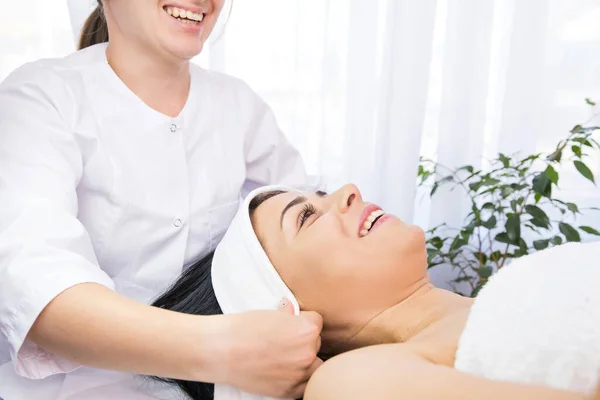 Cosmetologist rindo colocando toalha de cabeça branca na cabeça da mulher paciente para os procedimentos de rejuvenescimento do rosto em uma clínica de beleza. — Fotografia de Stock