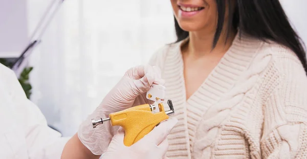 Fachärztin für medizinische Handschuhe hält Piercing-Pistole und medizinische Ohrringe in den Händen, die einer Kundin im Kosmetikschrank gezeigt werden. — Stockfoto