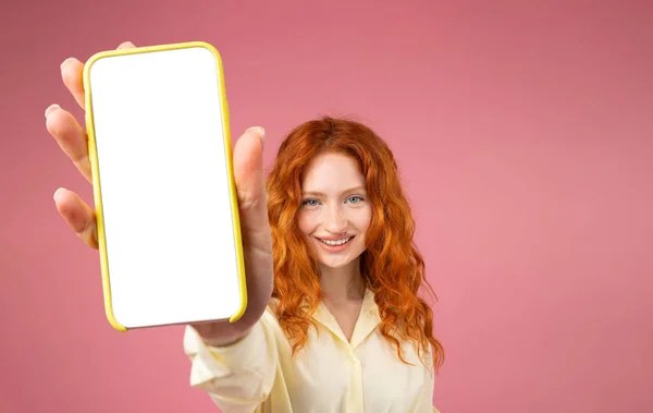 Porträtt av upphetsad rödhårig flicka som håller stor smartphone med vit blank skärm i handen, visar nära kameran. Gadget med tomt ledigt utrymme för mock up, banner isolerad på rosa bakgrund. — Stockfoto