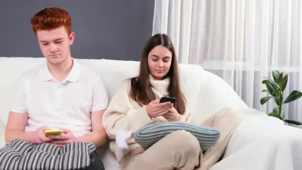 Młoda piękna para siedzi na kanapie w domu w luźnych ubraniach trzymając telefony komórkowe w rękach i patrząc na nie, gdy ktoś rzuca im papierowym samolotem.. — Wideo stockowe