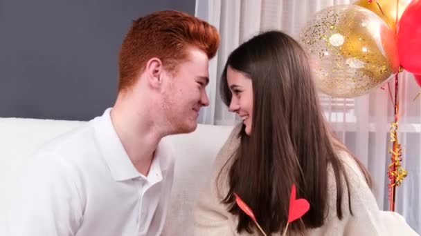 Amar pareja joven en casa riendo juntos mirándose, chica sosteniendo dos símbolos del día de San Valentín. — Vídeos de Stock