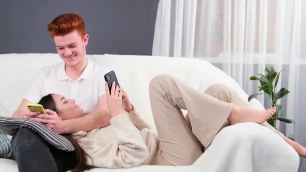 Wesoły rudzielec patrząc na telefon w jego rękach i pracując, jego dziewczyna położyła głowę na jego kolanie i ugięła kolana na kanapie.. — Wideo stockowe