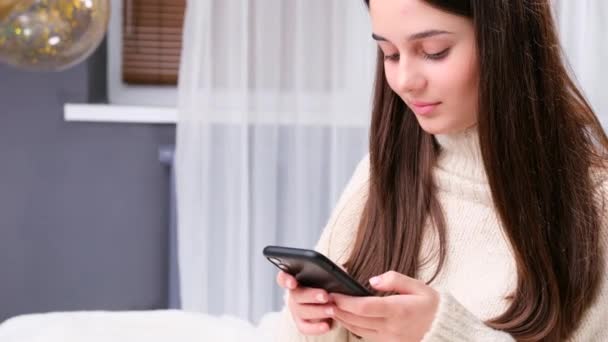 Glad smilende pige hjemme i hvid varm sweater ved hjælp af smart telefon til at chatte med sin kæreste, venter på at møde ham. – Stock-video