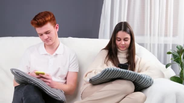 Videó egy fiatal párról, akik otthon ülnek párnákkal a térdükön, és mobiltelefonnal beszélgetnek.. — Stock videók