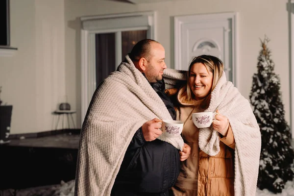 Прекрасна пара в зимовому одязі, покрита ковдрою на відкритому повітрі, тримає чашки гарячого чаю і посміхається разом . — стокове фото