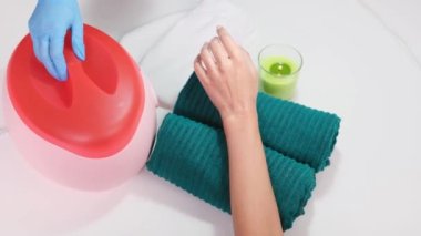 Havluya uzanmış bir kadın müşterinin videosunu kapat. Elinde bir bardak mum ve kozmetik uzmanı parafin banyosu yapmak için parafin kutusu açıyor..