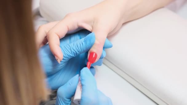 Maître professionnel en gants de protection bleus tenant la main de la femme et appliquant un vernis rouge de mode sur les ongles dans le salon de beauté. — Video