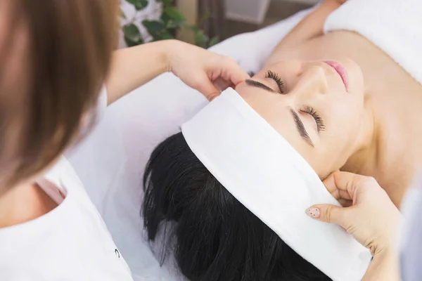 Cosmetologo professionista mettendo asciugamano bendaggio bianco ad una donna fronte per la terapia anti acne in clinica di bellezza. — Foto Stock